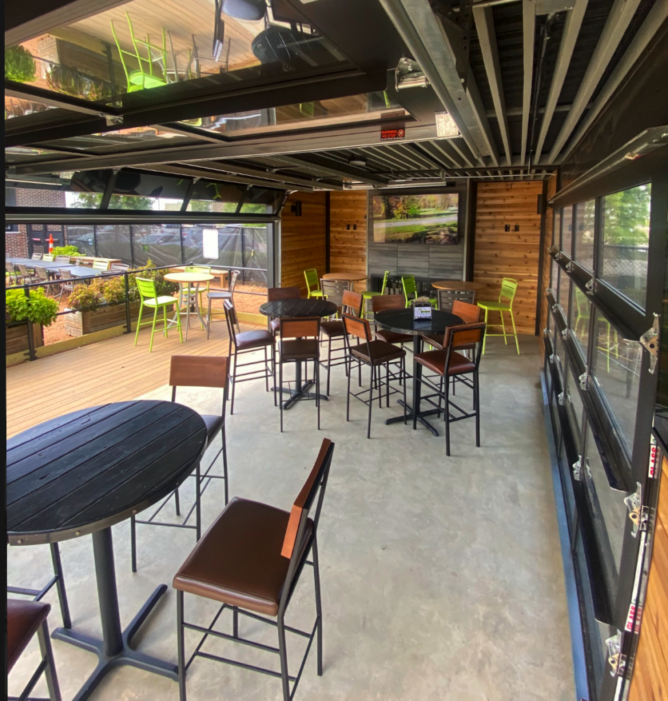 Modern indoor patio with glass garage doors, black round tables, brown chairs, and green chairs near planters. Sunlight streams in, and framed nature artwork hangs on the wooden wall in the background.